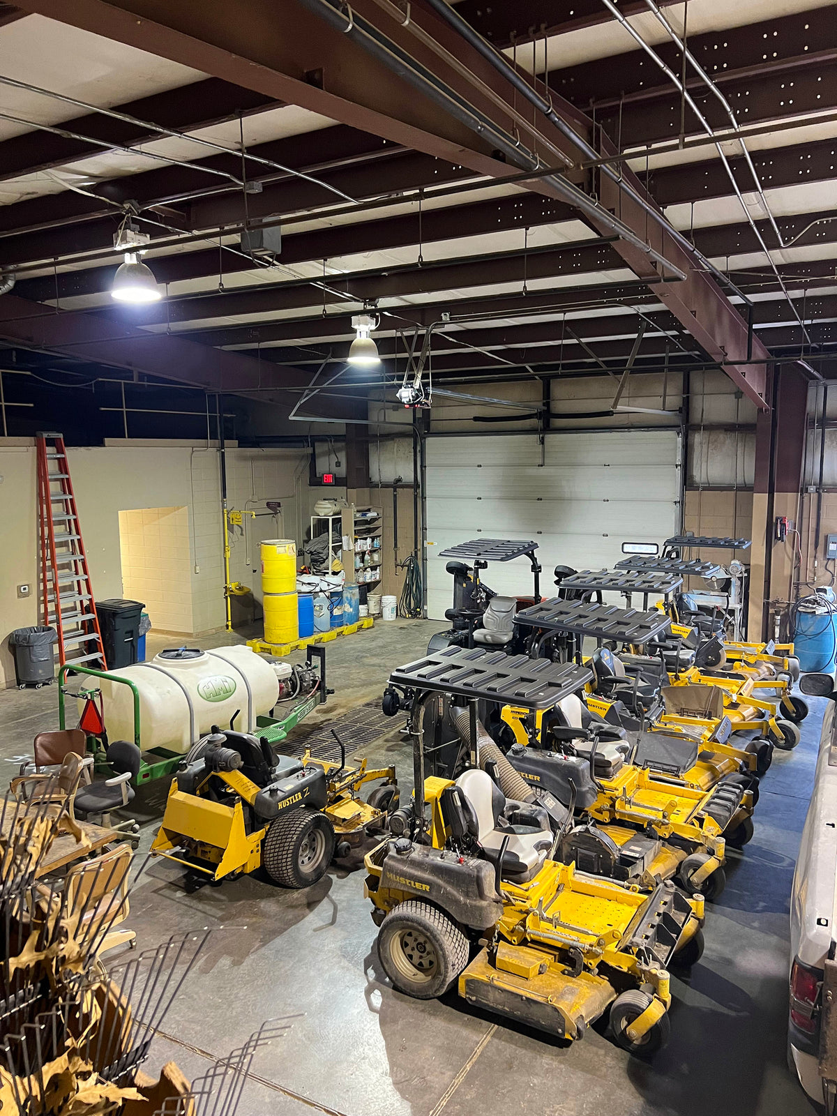 A large warehouse or farm equipment yard filled with yellow and black ride-on lawn mowers and other agricultural machinery, under industrial ceiling lights and exposed metal beams.