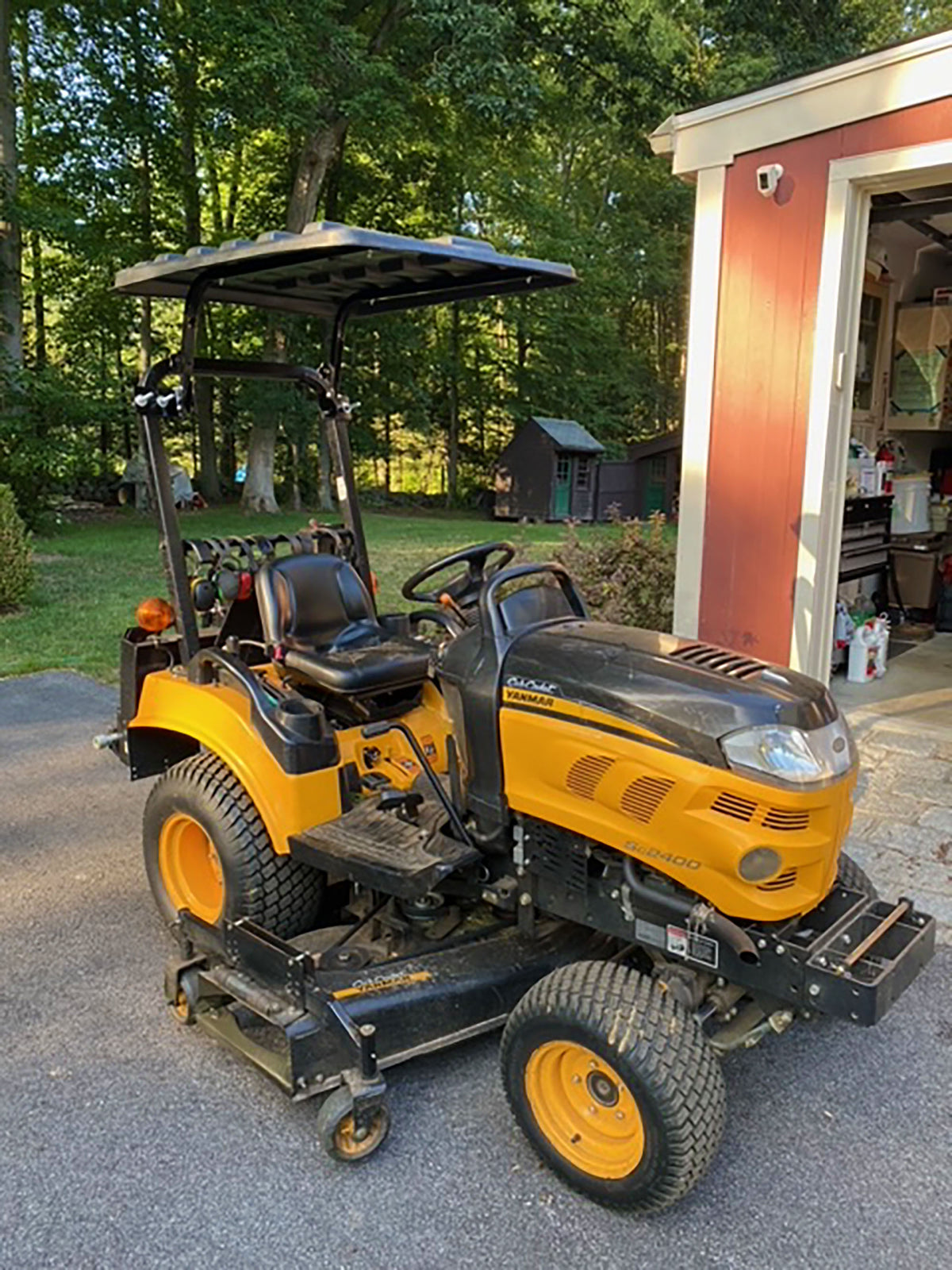 A yellow and black Kubota compact lawn tractor with a black canopy, parked on a driveway in front of a red garage door with trees in the background.