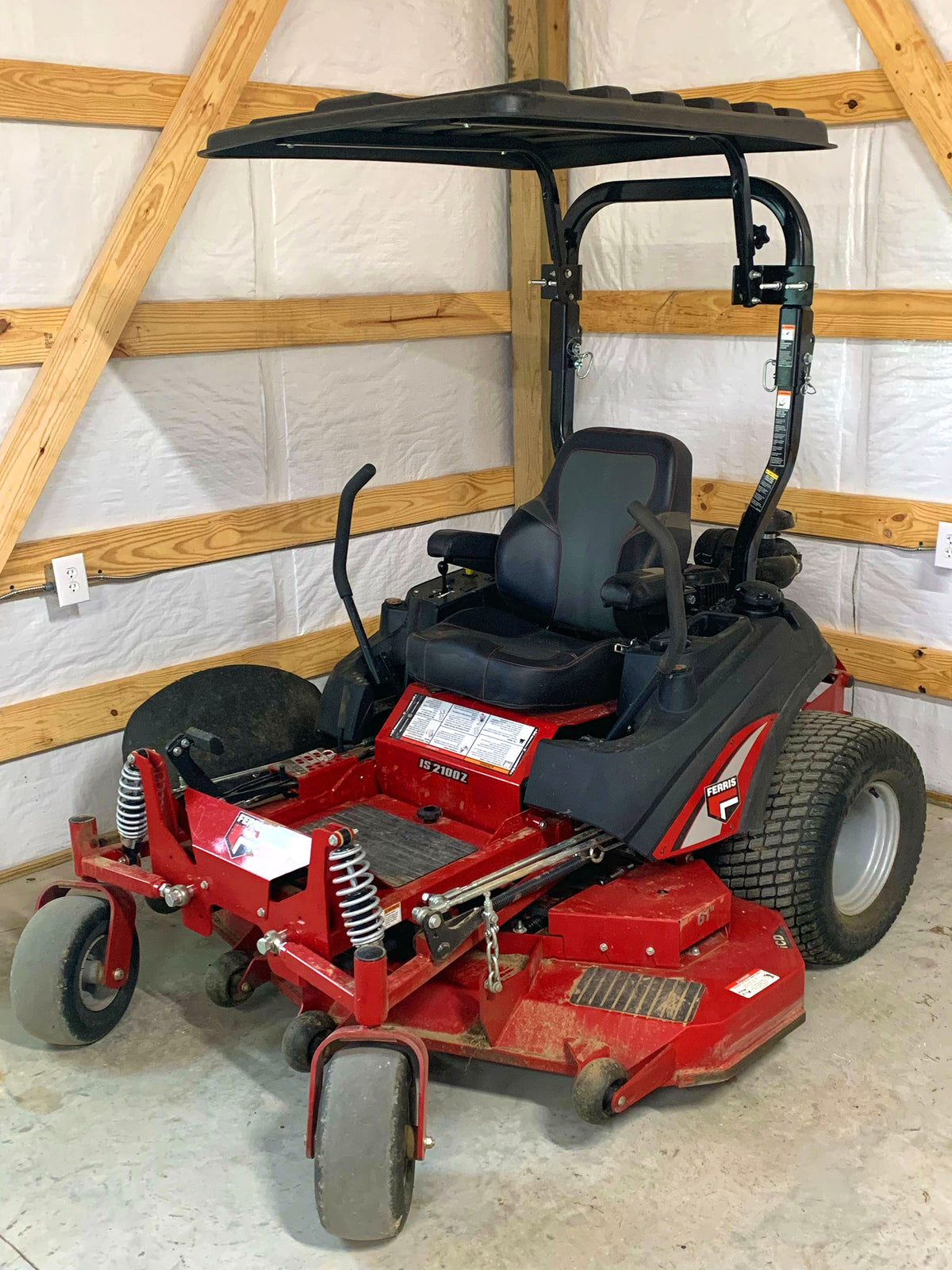 A red and black riding lawn mower with an overhead canopy, photographed inside a warehouse or garage with exposed wooden beams and white walls.