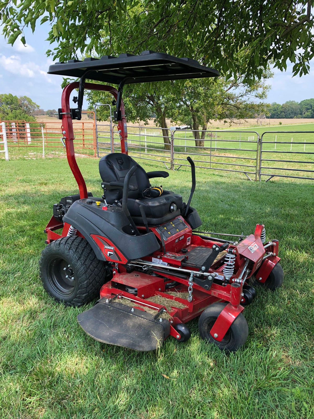 A red and black zero-till riding lawn mower with an enclosed driver seat and canopy, parked on a green grassy field with a fence and trees in the background.