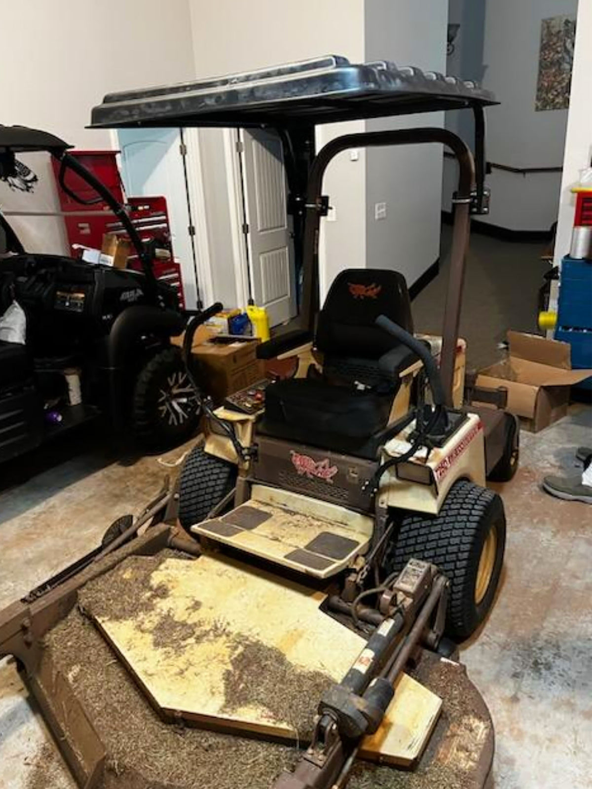 A used beige and black ride-on lawn mower with a canopy and front blade attachment, photographed in a garage setting.