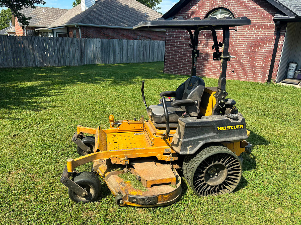 A yellow and black Hustler zero-till lawn mower with an overhead roll bar, parked on a green grass lawn in front of a brick house.