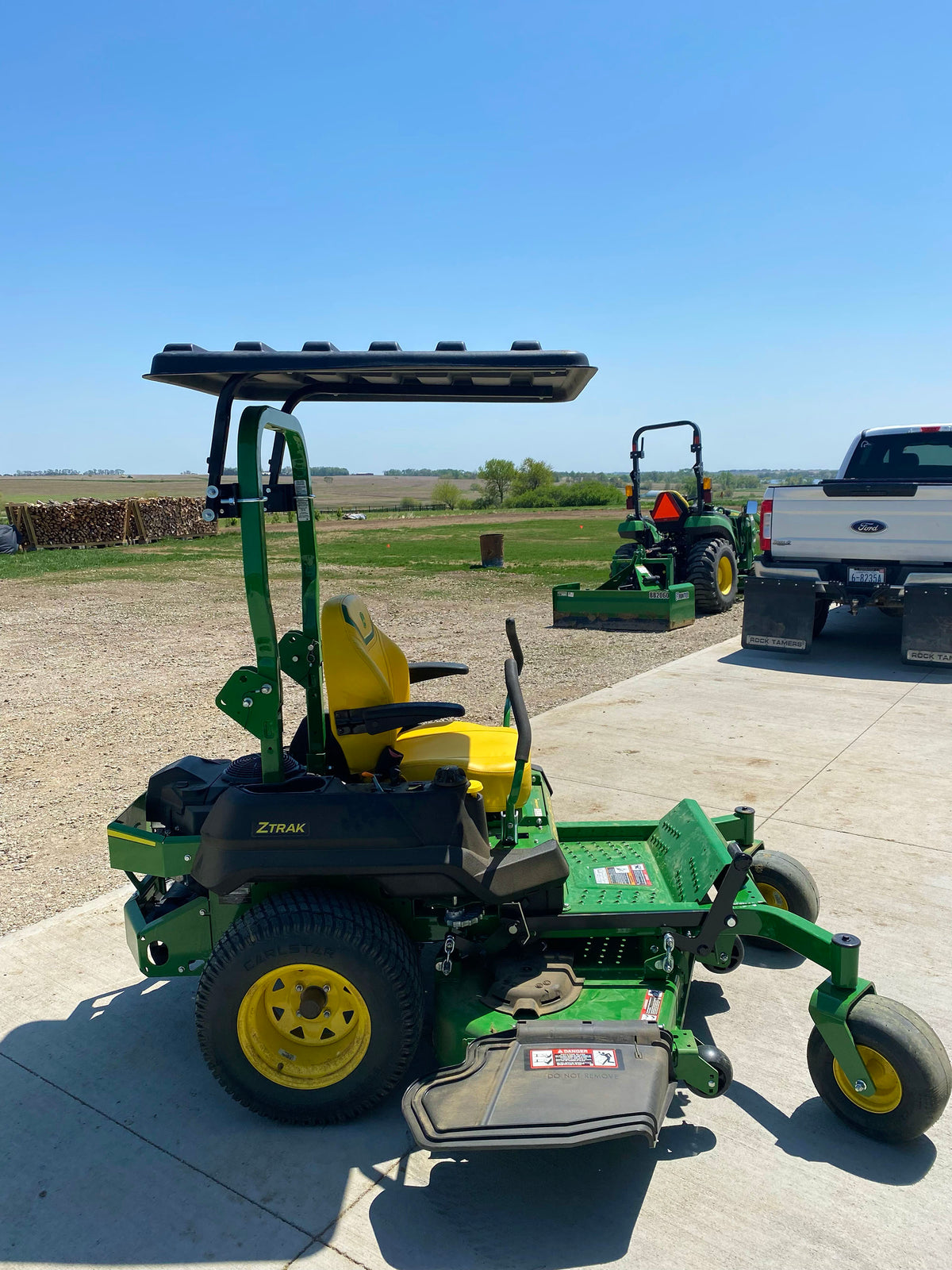 A green and yellow ZTAK zero-till riding lawn mower with a black canopy, parked on a concrete driveway outdoors. A white pickup truck and another tractor are visible in the background under a clear blue sky.