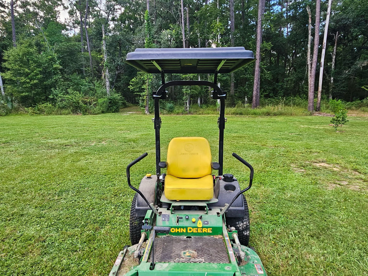 A John Deere zero-till lawn mower with a yellow seat and a black canopy roof, positioned on a green grass lawn with a forest background.