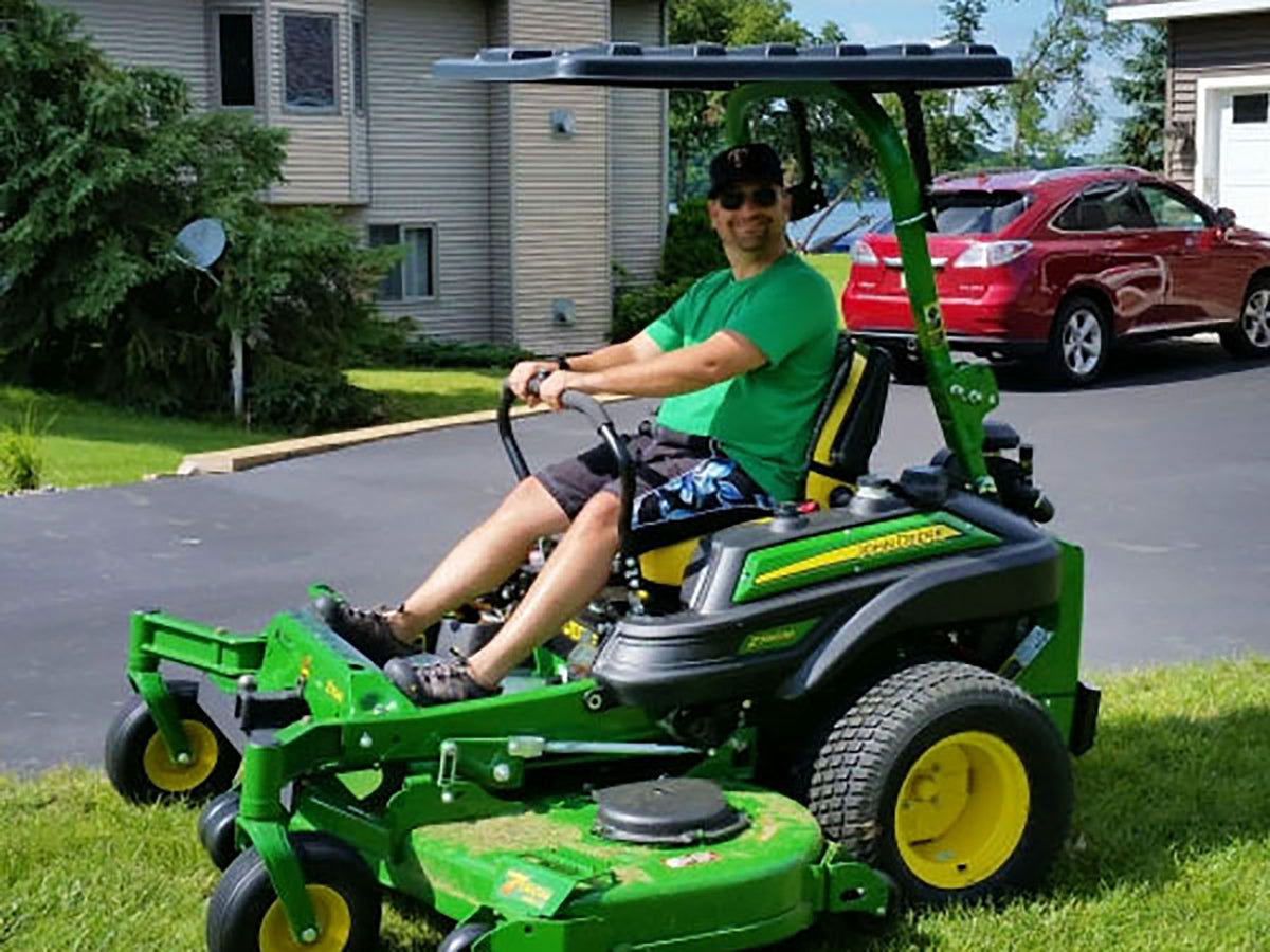A man wearing a green t-shirt and sunglasses is driving a green and black riding lawn mower with yellow wheels on a grassy lawn. A red SUV is parked in the background on a paved driveway next to a house.