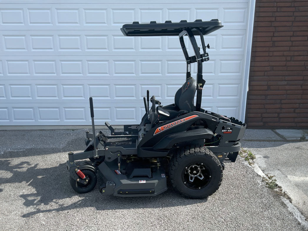 A black riding lawn mower with an open canopy and rear deck, parked on asphalt in front of a white garage door and brick wall.