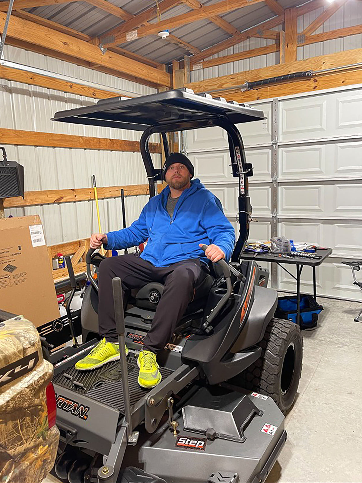A man wearing a blue hoodie, black beanie, and neon yellow sneakers sits in the driver's seat of a gray step-on ride-on lawn mower inside a garage.